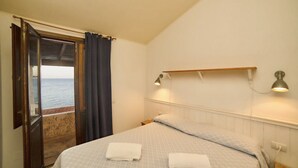 A bedroom with a bed, two bedside lamps, a nightstand, a window with curtains, and a view of the sea.