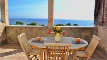 Une table ronde en bois, entourée de quatre chaises, un vase rempli de fleurs et deux tasses orange, le tout posé sur une terrasse dallée de pierres, d’où l’on a une vue sur la mer.