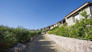 A paved pathway flanked by stone walls and greenery, leading to a row of traditional buildings with terracotta roofs.