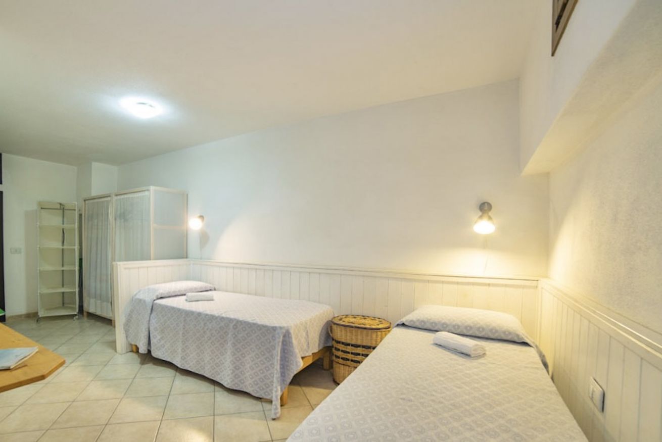 Una habitación con dos camas individuales, una mesa de madera, una cesta de mimbre y un armario blanco.