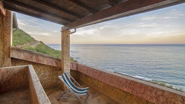 A covered outdoor area with a view of the sea, a stone bench, and a striped beach chair.