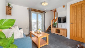A living room with a blue sofa, wooden coffee table, and a TV on a wooden stand. A staircase leads to a balcony with a view. - Roosters Rest - UK48255 (Downderry)