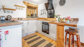 A modern kitchen with white cabinets, a wooden countertop, a microwave, and a kettle. There is a wooden shelf above the sink, a clock on the wall, and a fire extinguisher on the wall. - Roosters Rest - UK48255 (Downderry)