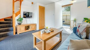 A modern living room with a wooden staircase, a TV mounted on the wall, a wooden coffee table with cups and plates, and a blue sofa with colorful cushions. - Roosters Rest - UK48255 (Downderry)