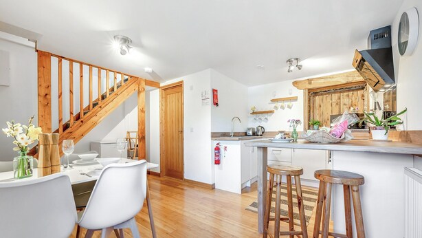 A modern kitchen with wooden accents, a dining area with white chairs, and a staircase. - Roosters Rest - UK48255 (Downderry)