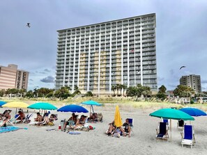 A beach with rows of beach chairs and umbrellas, a tall building in the background, and a clear sky.