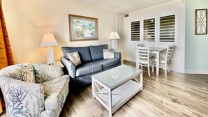 A living room with a grey sofa, a glass coffee table, and a white dining table with chairs. There are two lamps and a framed picture on the wall. - Tropical Resort 1BR 307 (North Myrtle Beach)