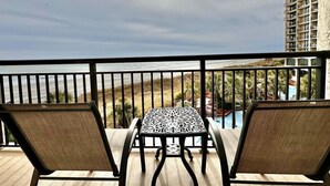 A balcony with two wicker chairs and a table, overlooking a beach and a pool area. - Tropical Resort 1BR 307 (North Myrtle Beach)