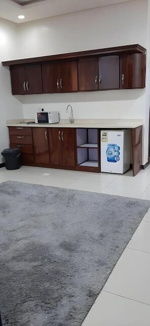 A compact kitchen with dark wooden cabinets, a white countertop, a microwave, and a refrigerator. - Ahlin Suites 3 (Riyadh)