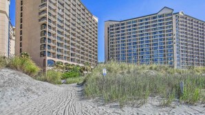 A beachfront hotel with multiple high-rise buildings, sandy beach, and sparse vegetation. - Tropical Resort 1BR 1407 (North Myrtle Beach)