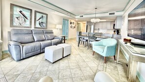 A living room with a gray sofa, ottoman, and a blue armchair. There is a dining area with a table and chairs, and a kitchen area with cabinets and a TV.