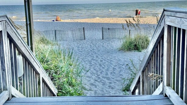 Ein hölzerner Steg führt zu einem Sandstrand mit freiem Blick auf das Meer, eine Person ist in der Ferne zu sehen.