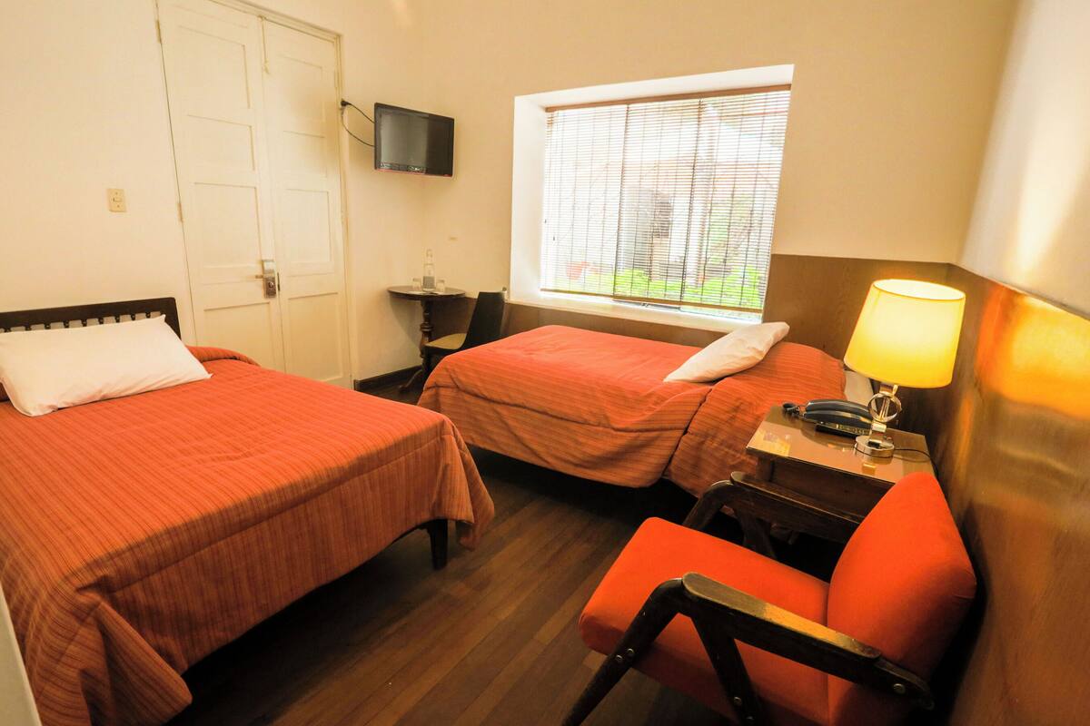 A room with two beds, a desk with a lamp, a chair, a TV on the wall, and a window with blinds.