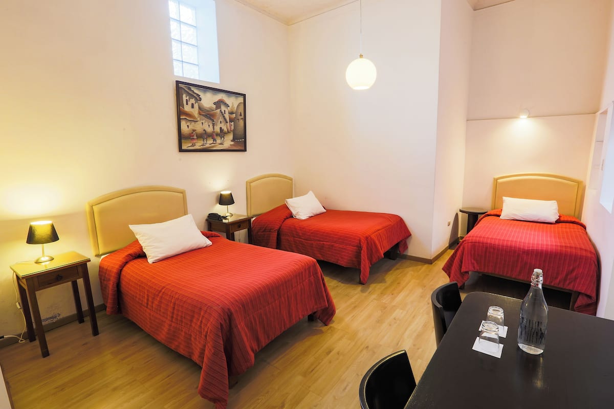 A hotel room with three beds, a table with a bottle, and a painting on the wall.