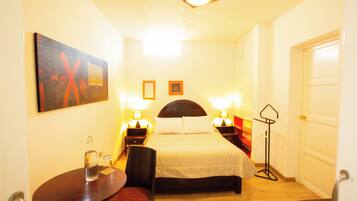 Habitación de hotel con cama, mesitas de noche con lámparas, una silla, una mesa con una botella y un cuadro en la pared.