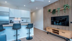 A modern kitchen with white cabinets, a stainless steel refrigerator, and a built-in oven. There are three black bar stools and a wooden dining table with chairs. A flat-screen TV is mounted on a wooden wall with plants.