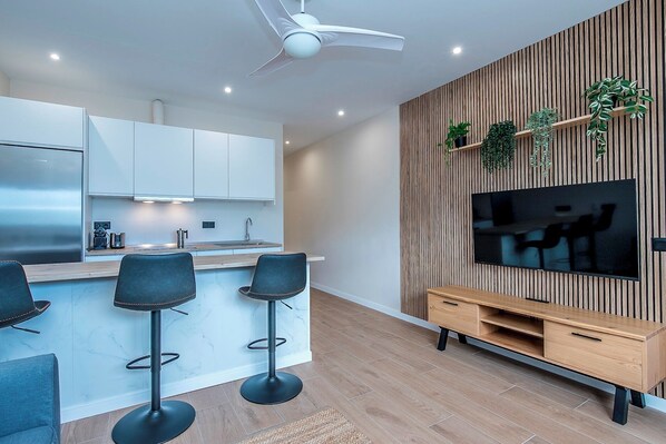 A modern kitchen with white cabinets, a stainless steel refrigerator, and a built-in oven. There are three black bar stools and a wooden dining table with chairs. A flat-screen TV is mounted on a wooden wall with plants.