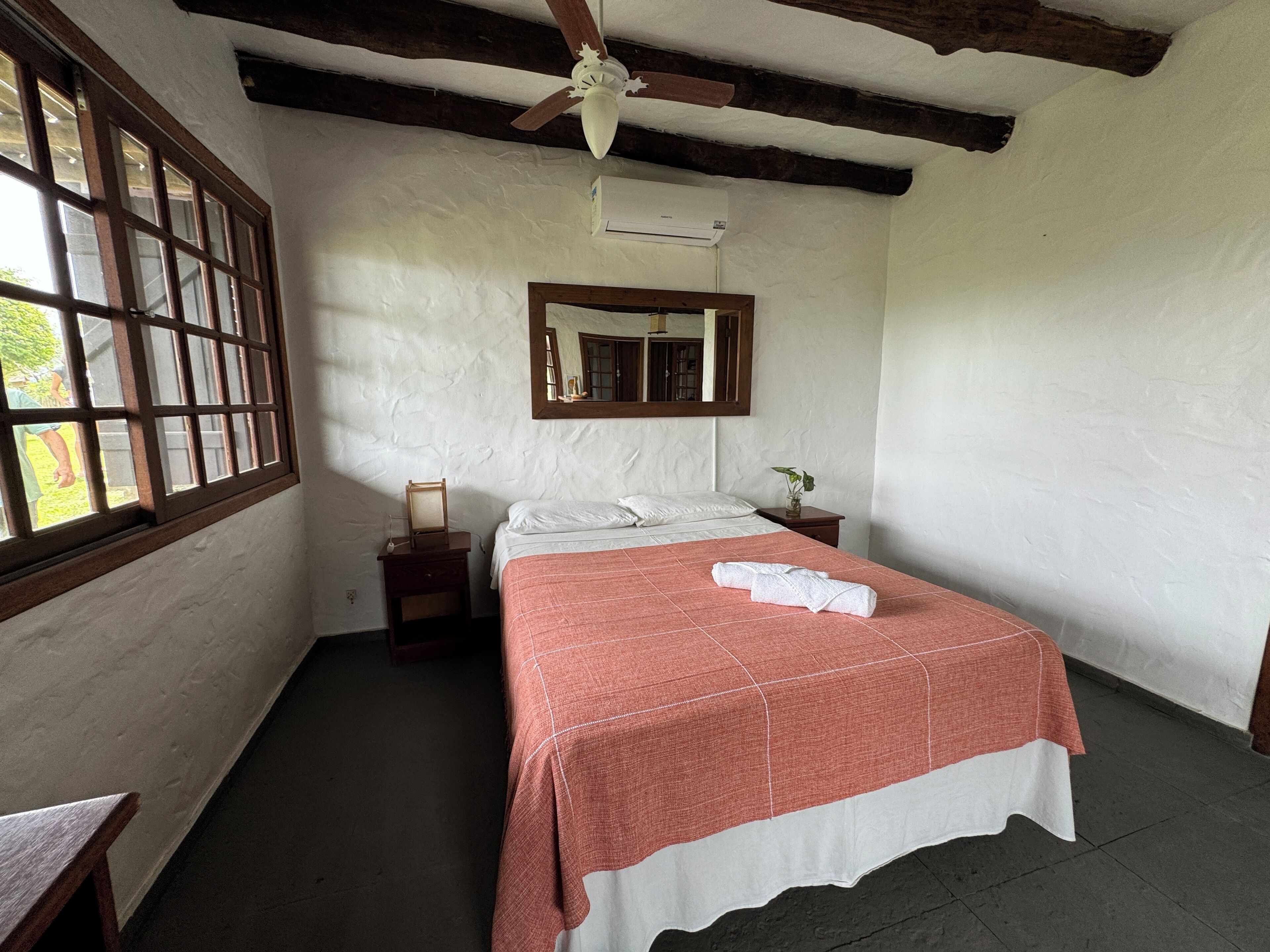 Un dormitorio con cama, ventilador de techo, ventana, espejo y mesita de noche.