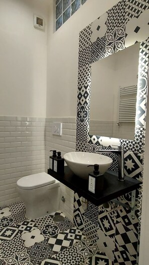 Un baño con inodoro blanco, lavamanos negro y espejo. Las paredes presentan un diseño de azulejos con estampado blanco y negro.