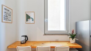 Une petite cuisine avec une table en bois, deux chaises blanches, une bouilloire, une plante en pot et une fenêtre donnant sur un beau paysage.