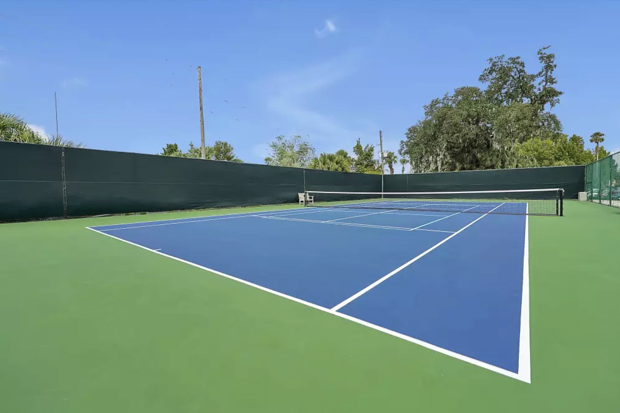 Een tennisbaan met een net, omringd door een groene hekken en bomen.