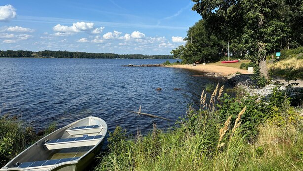 A calm lake with a small boat docked at the shore, surrounded by greenery and a sandy beach. - 3 bedroom gorgeous home in Ljungby (Ljungby)