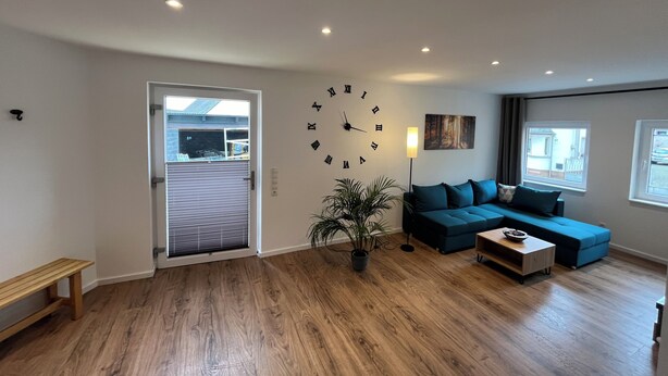 A spacious living room with a large wall clock, a blue sofa, a wooden coffee table, and a potted plant.