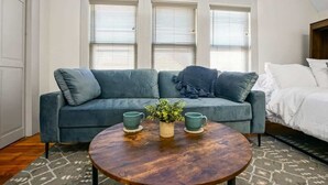 Ein Zimmer mit einem blauen Sofa, einem runden Holztisch mit zwei grünen Tassen und einer kleinen Pflanze sowie einem Bett mit weißer Bettwäsche.