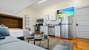 Ein kompaktes Hotelzimmer mit Bett, Sofa, Esstisch mit blauen Tellern, einer Küche mit modernen Geräten und einem großen Fernseher, der eine Strandansicht zeigt.