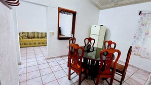 Un coin repas avec une table en verre et des chaises en bois, un réfrigérateur blanc et un canapé à l’arrière-plan.