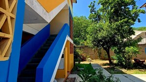 Un bâtiment à la façade bleue et jaune, avec un escalier et un jardin agrémenté d’un arbre et de plantes.