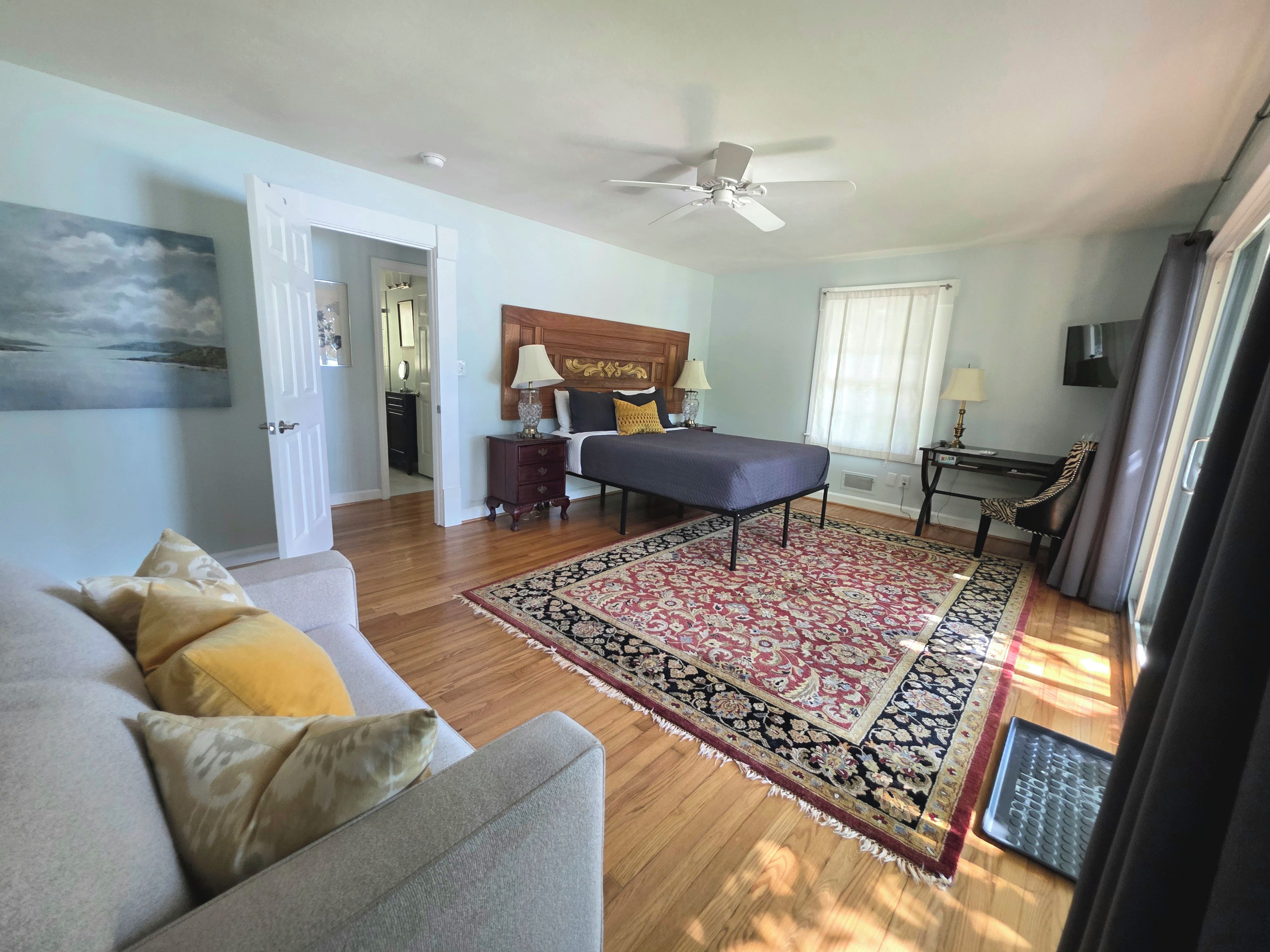 A bedroom with a bed, bedside tables, a ceiling fan, a window with curtains, and a view of the hallway.