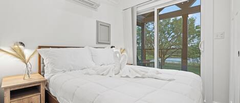 Un dormitorio con una cama grande, una mesita de noche, una ventana con vista a árboles y un ventilador de techo.