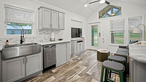 A modern kitchen with white cabinets, a stainless steel sink, and a dishwasher. There is a ceiling fan, a large window with blinds, and a view of the outdoors.