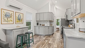 A modern kitchen with grey cabinets, a marble countertop, and a herringbone wood floor. There are two green stools and a window with blinds.
