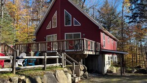 A red cabin with a wooden deck and stairs leading to a covered porch. - Adirondack Red House, Adirondack Red House (Eagle Bay)