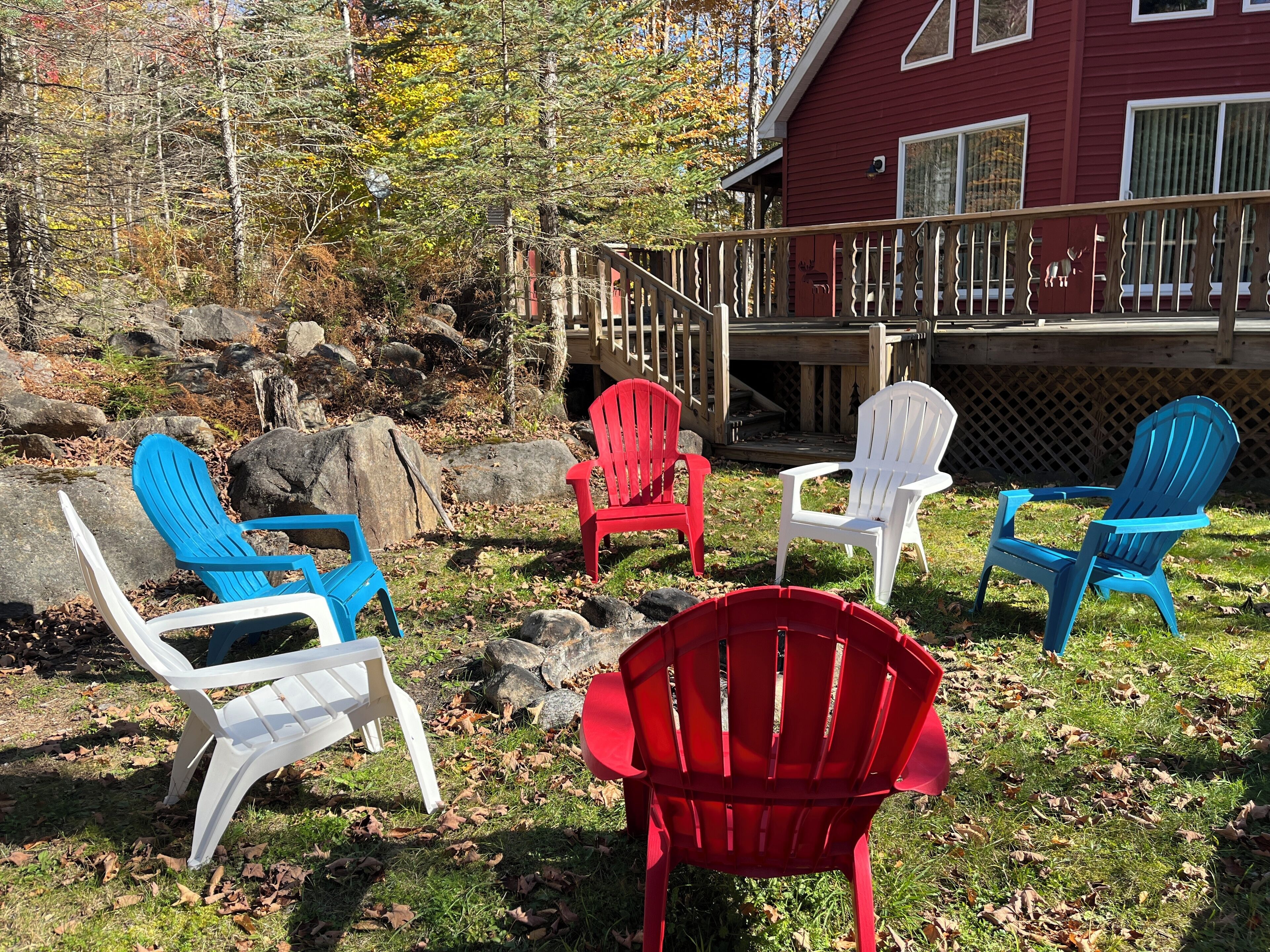 En træterrasse med Adirondack-stole i rødt, hvidt og blåt, omgivet af et naturlandskab med træer og sten.
