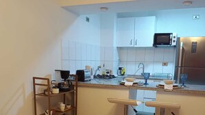 Et moderne køkken med hvide skabe, et rustfrit stål køleskab, en mikrobølgeovn og en kaffemaskine. To barstole og en lille hylde med planter.