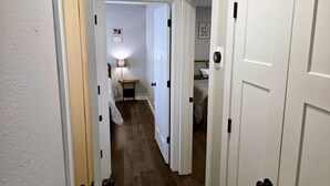 A narrow hallway with white doors, a small bedside table with a lamp, and a bed with white bedding.