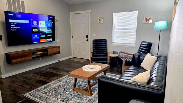 A living room with a flat-screen TV, a black leather sofa, a wooden coffee table, a patterned rug, and a door.