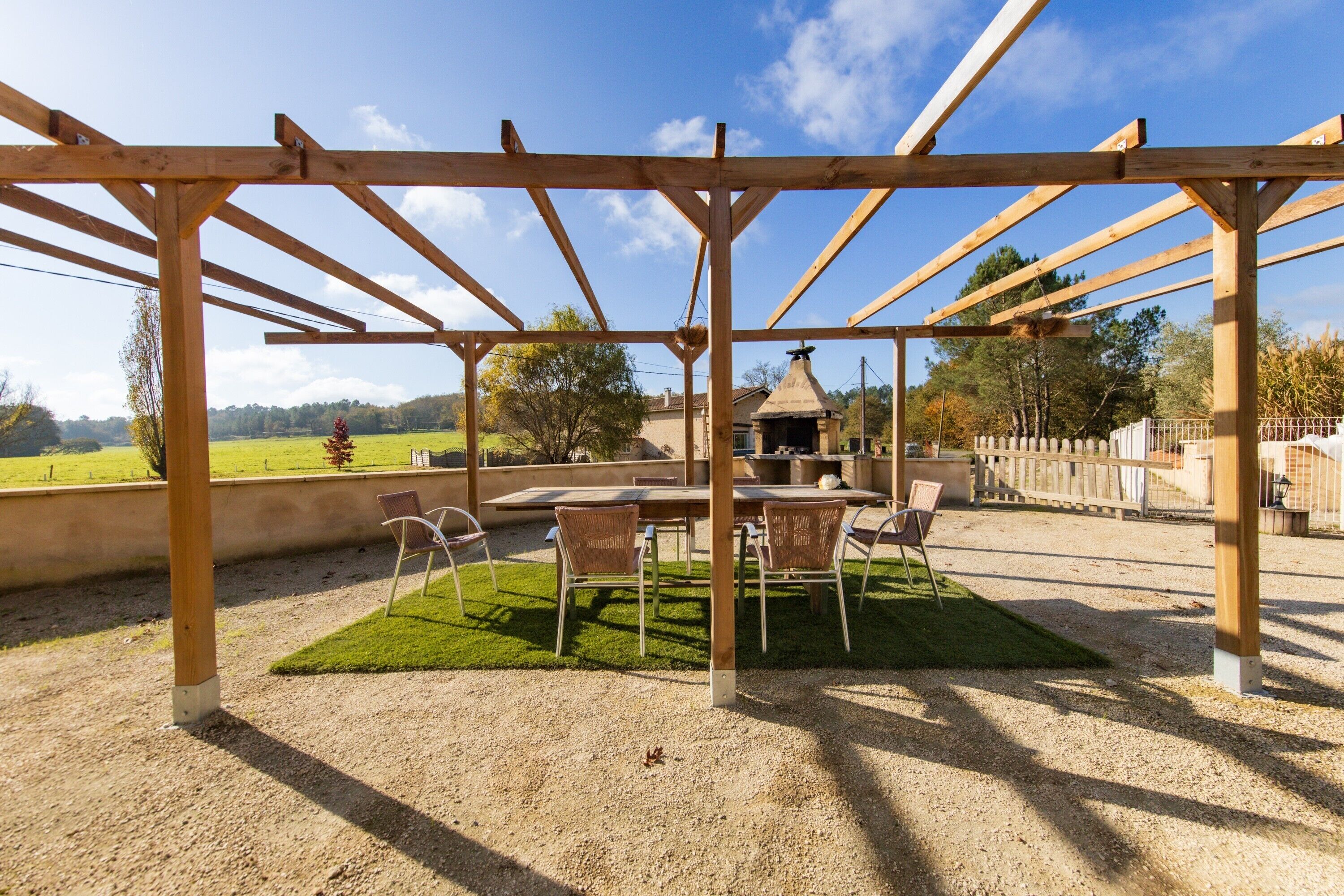Une structure en bois formant une pergola, avec une table et des chaises en dessous, entourée d’une zone recouverte de gravier et d’une petite pelouse.