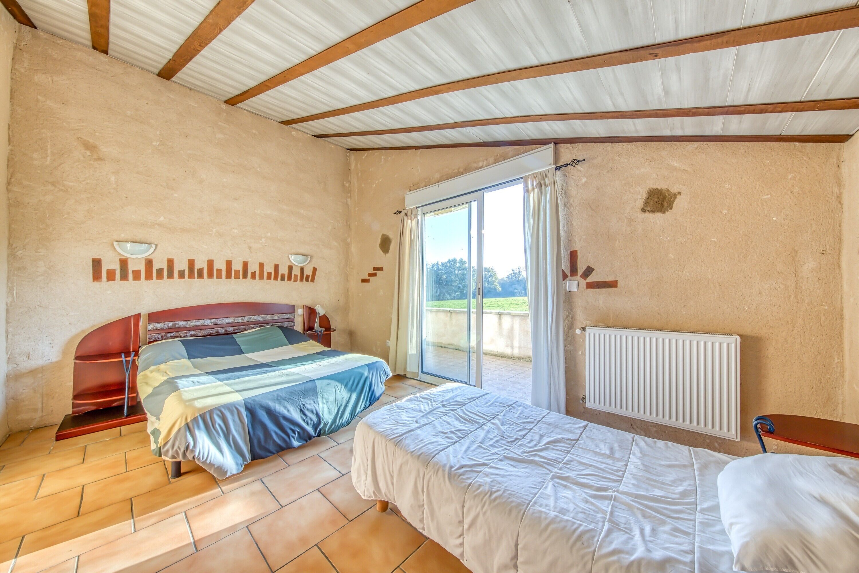 Quarto com duas camas, porta de correr de vidro para uma varanda, radiador e estrutura de teto em madeira.