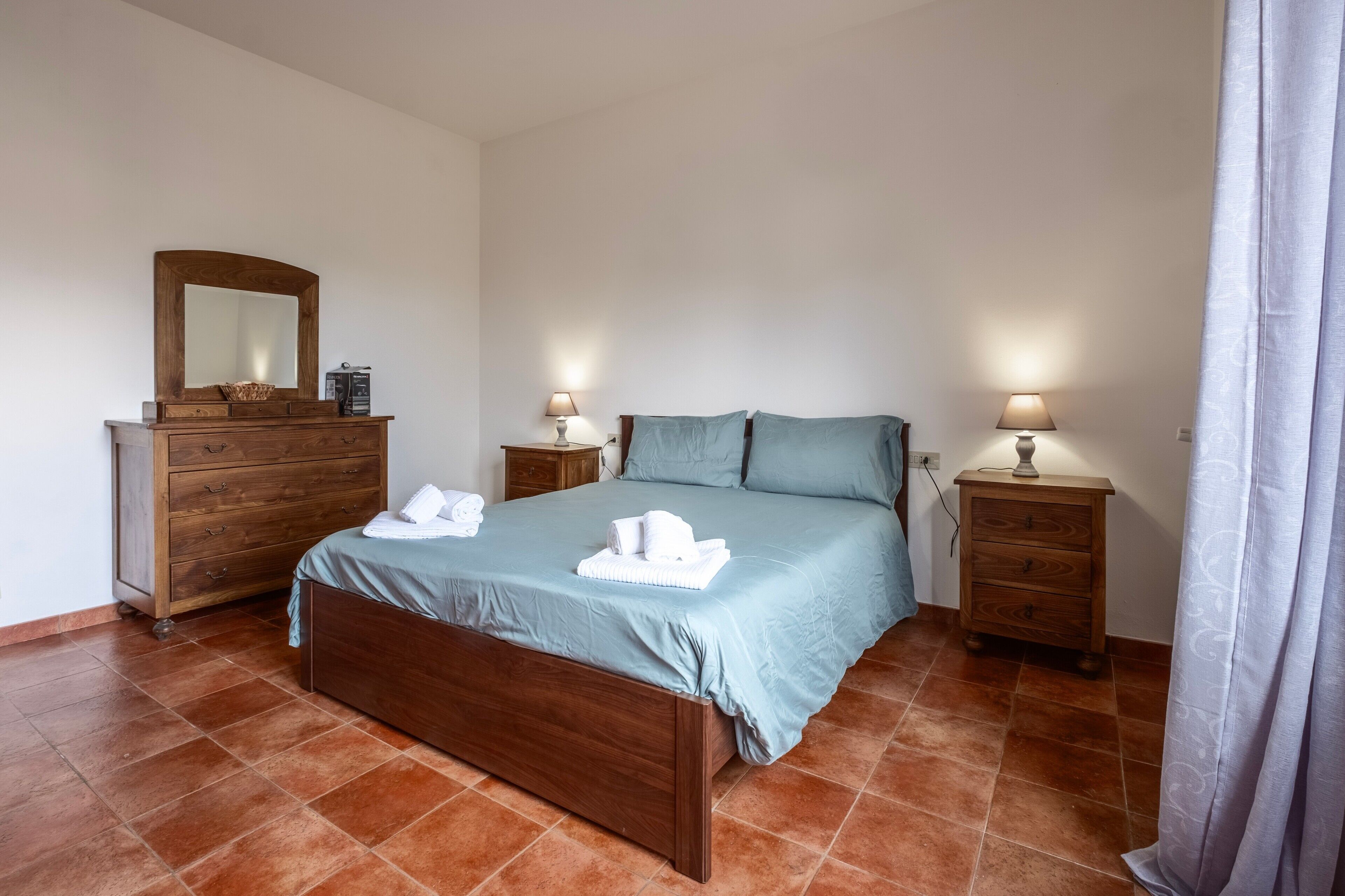 Un dormitorio con cama, cómoda de madera, dos mesitas de noche con lámparas y un espejo.