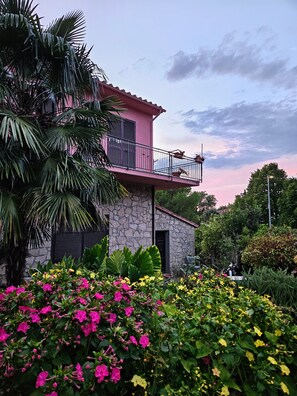 A two-story building with a balcony, surrounded by lush greenery and flowering plants.
