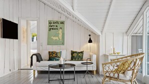 Une pièce chaleureuse avec un plancher en bois blanc, un canapé blanc agrémenté de coussins à motifs floraux, un fauteuil en osier, une petite table ronde avec une bougie et une affiche murale.