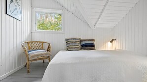Une chambre avec un grand lit, un fauteuil en osier, deux coussins à motifs, une fenêtre donnant sur des arbres et une lampe fixée au mur.