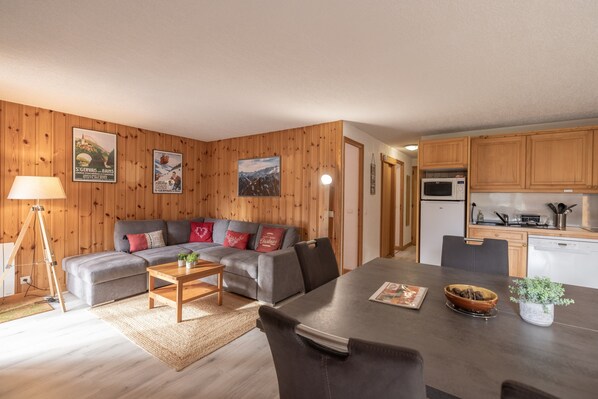A living room with a sofa, coffee table, and a dining table with chairs. There is a kitchen area with wooden cabinets and appliances.