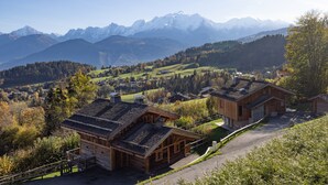 Een houten chalet met een schuin dak, een stenen fundering en uitzicht op een vallei met bergen op de achtergrond.