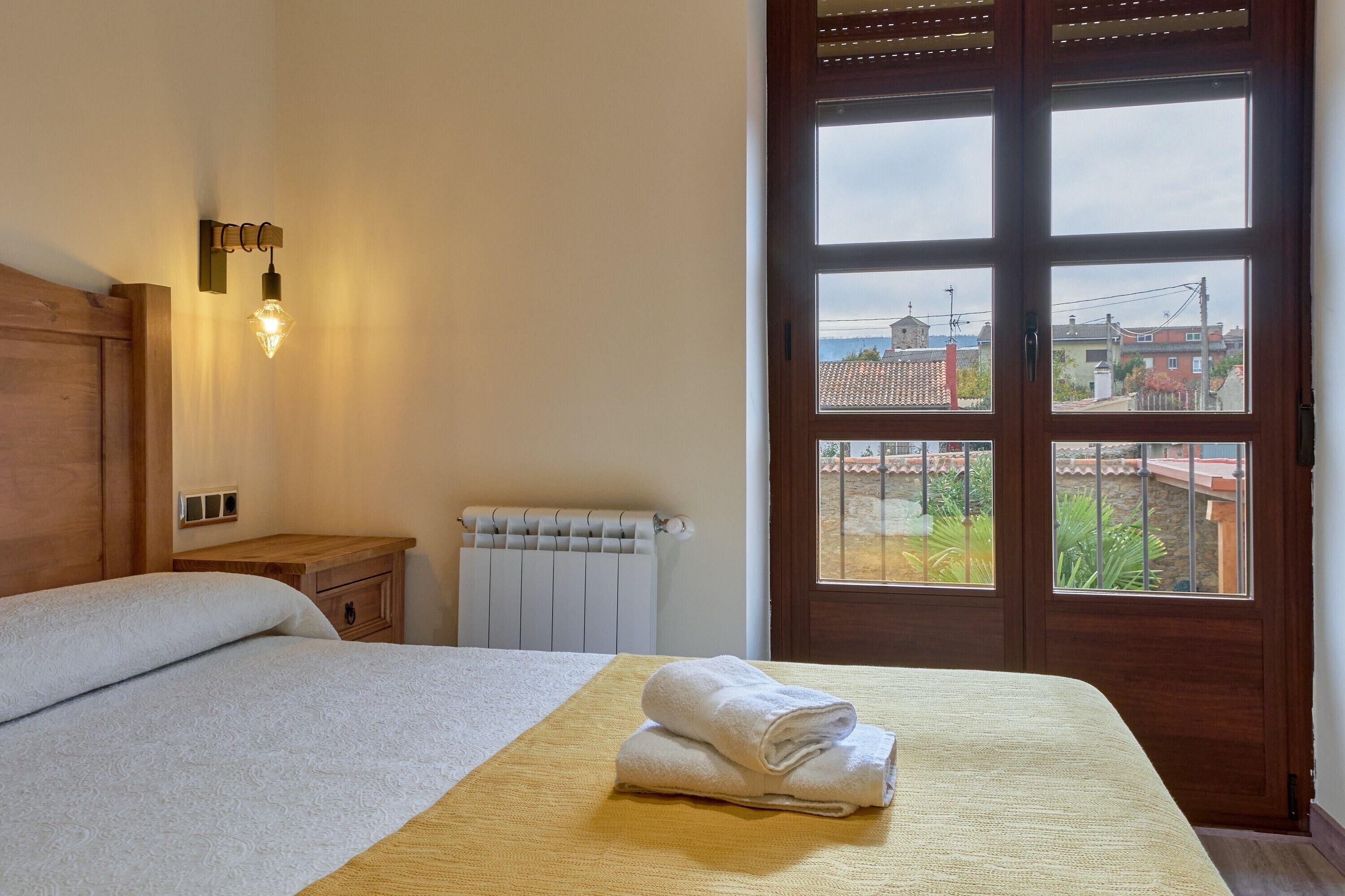 Une chambre à coucher avec un lit, une table de chevet, un radiateur et une vue sur l’extérieur, visible à travers une porte en bois.