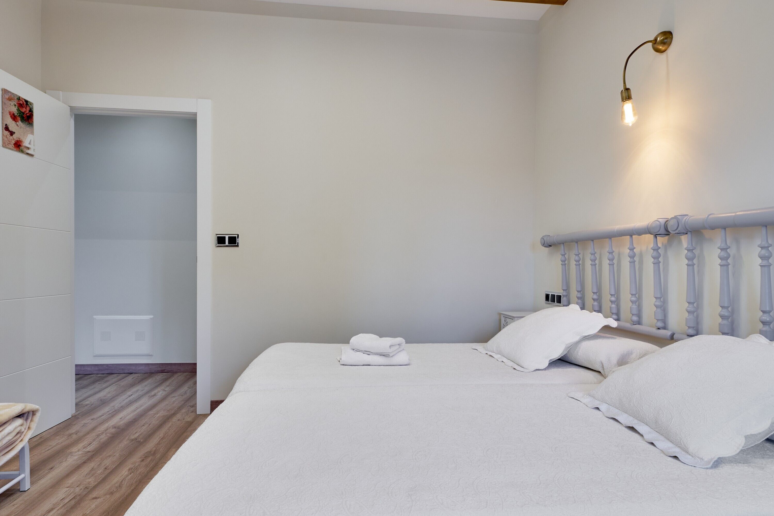 A bedroom with a bed, white bedding, a nightstand, a wall-mounted light, and a door leading to another room.